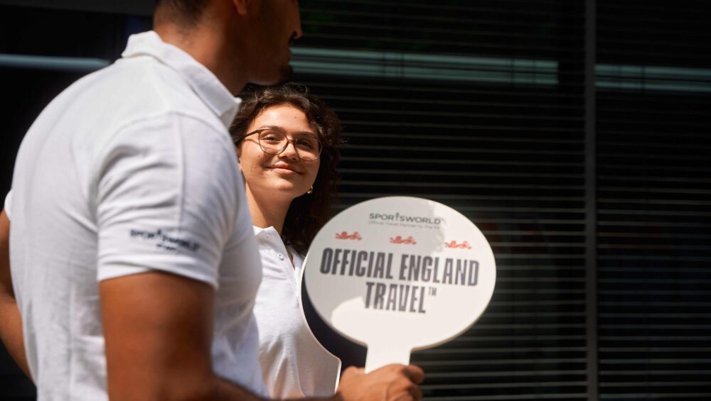 Two Sportsworld employees hold a sign saying 'Official England Travel' while welcoming fans at UEFA Euro 2024