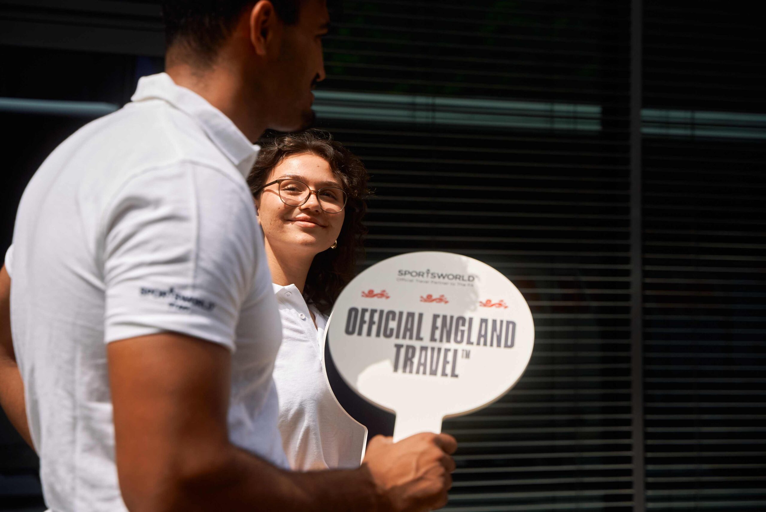 Two Sportsworld employees hold a sign saying 'Official England Travel' while welcoming fans at UEFA Euro 2024